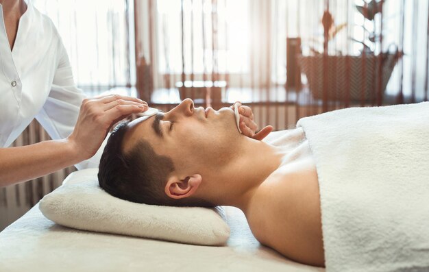 Cosmétologue femme nettoyant le visage de l'homme avec deux éponges dans le salon spa, vue latérale