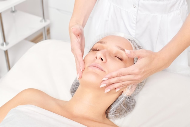 Le cosmétologue masse le masque crème sur la peau du visage de la femme pour une procédure de rajeunissement dans un salon de beauté