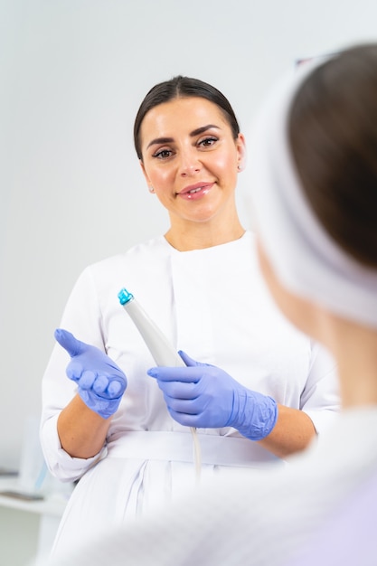 Cosmétologue souriante dans des gants en latex avec un appareil de microdermabrasion à la main en regardant son patient