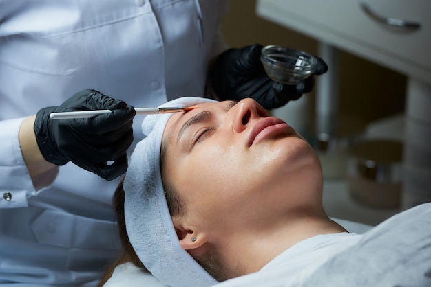 la cosmétologue utilise une brosse pour appliquer un peeling transparent sur le visage de la femme.