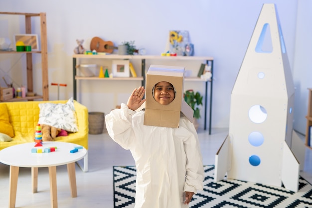 Cosmonaute drôle mignon en vêtements blancs et boîte en carton sur sa tête en gardant la main droite par le temple tout en jouant dans le salon