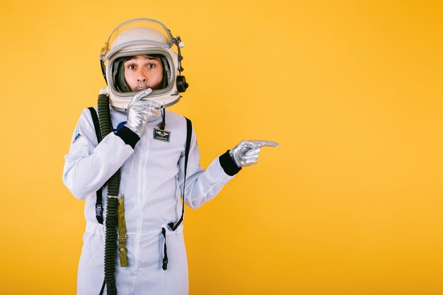 Cosmonaute masculin surpris en combinaison spatiale et casque, pointant le doigt vers la droite, sur le mur jaune.