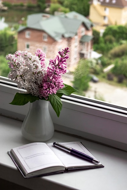 Cosy home still life spring lilas flowers in a white vase on the windowill notebook and glasses Retro style