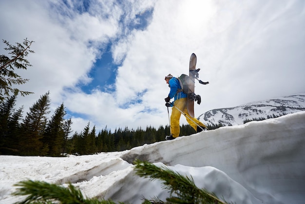 Photo côté du snowboarder face cachée avec snowboard sur le dos sur une pente enneigée nuages sur ciel bleu sur fond angle bas