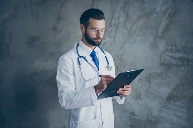 Côté profil photo de l'homme chirurgien concentré entendre les plaintes des patients écrire dans son presse-papiers porter un manteau médical blanc isolé sur un mur de couleur grise