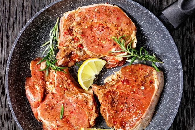 Côtelettes de porc épicées crues avec des brins de romarin et des tranches de citron dans une poêle sur une table en bois, vue horizontale d'en haut, mise à plat, gros plan