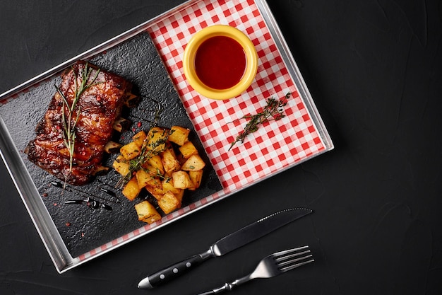 Côtes levées au barbecue avec pommes de terre au four et sauce chili piquante sur un plateau en fer. Fond noir