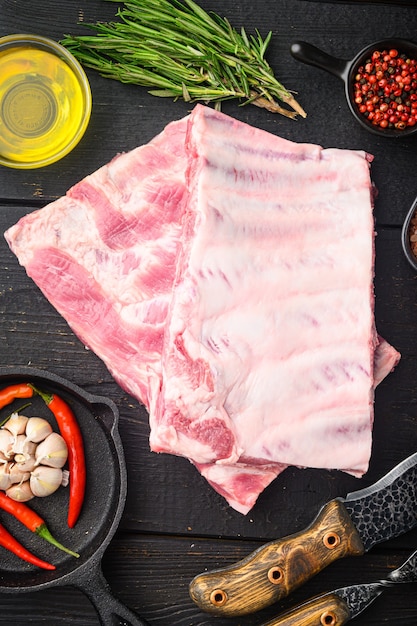 Côtes de porc crues fraîches avec du romarin et de l'ail, avec un couteau à barbecue et une fourchette à viande, sur une table en bois noir, vue de dessus à plat