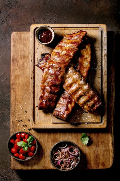 Côtes de porc grillées au barbecue servies avec des oignons marinés, des tomates cerises, du basilic et de la sauce barbecue sur une planche à découper en bois sur une surface texturée brun foncé. Vue de dessus, mise à plat