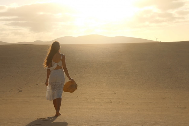 Coucher de soleil dans le désert. Jeune femme en robe blanche marchant dans les dunes du désert avec des pas dans le sable pendant le coucher du soleil. Fille qui marche sur le sable doré sur Corralejo Dunas, Fuerteventura.