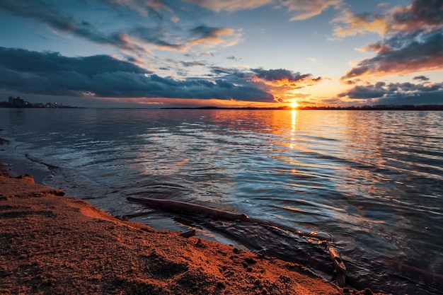 Coucher de soleil sur la rive du fleuve. De beaux nuages, de l'eau bleue pétillante, une branche d'arbre tombée dans l'eau et du sable sur la rive orange des rayons du coucher du soleil.