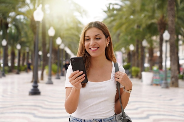 Coup moyen jeune femme souriante utilise un smartphone lit les nouvelles des réseaux sociaux recherche le texte envoie des mails en ligne se promène dans la rue Mode de vie technologique des gens