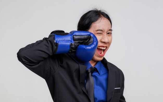 Coup de studio isolé d'un stress fou asiatique déprimant un homme d'affaires fou en costume formel noir debout portant un gant de boxe bleu frappant et blessant son propre visage en criant de douleur sur fond blanc.