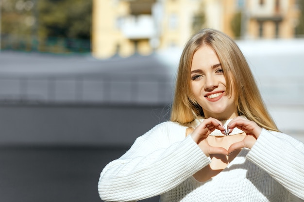 Coup de style de vie : magnifique fille blonde en pull tricoté blanc faisant un symbole de coeur avec ses mains. Espace pour le texte