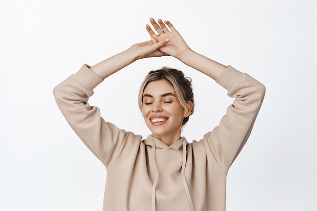 Coup de taille d'une femme blonde heureuse, levant les mains en se sentant libre et joyeuse, souriant les yeux fermés, dansant sur blanc.