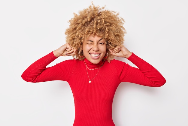 Coup de taille d'une femme bouclée joyeuse ignore la musique forte, bouche les oreilles avec les doigts, cligne des yeux et sourit à pleines dents, porte un col roulé rouge décontracté ne peut pas supporter un bruit fort isolé sur fond blanc