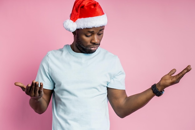 Coup de taille d'un homme afro-américain abasourdi écartant les mains, portant un bonnet de Noel, un t-shirt, regardant perplexe, ne comprenant pas ce qui s'est passé, isolé sur fond rose.