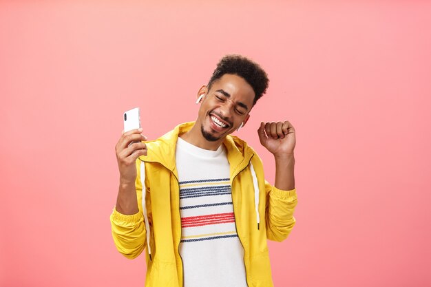 Coup de taille d'un homme afro-américain optimiste et insouciant dans des écouteurs sans fil tenant un smar...