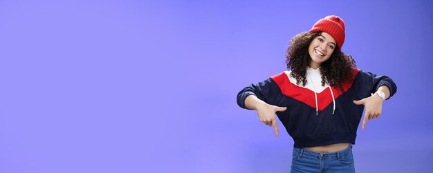 Photo coup de taille d'une jeune femme cool et élégante aux cheveux bouclés souriante et joyeuse pointant vers le bas