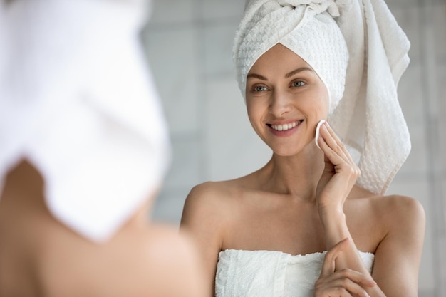 Coup de tête gros plan réflexion miroir souriante belle femme enveloppée dans une serviette appliquant du toner avec un coton sur la peau visage après le bain Heureuse jeune femme faisant la routine de soins de la peau du matin après la douche