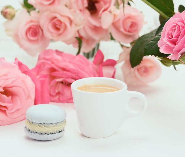 Photo coupe en céramique blanche avec du café et du macaron bleu sur une table blanche derrière un bouquet de roses roses