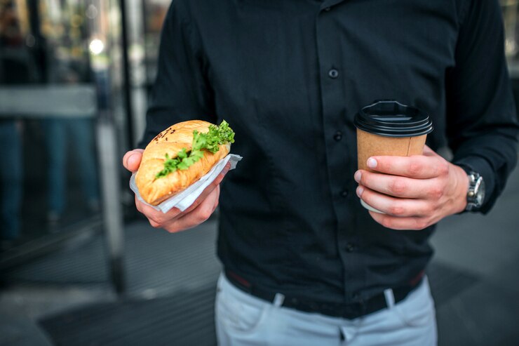 Photo coupe de l'homme tenant un croissan salé et une tasse de boisson. il se tient à la construction. guy porte une chemise noire et un jean bleu.