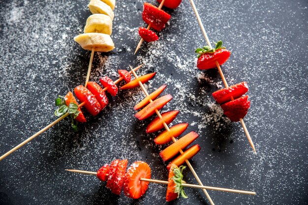 Couper les fraises et les bananes aux prunes sur une brochette avec du sucre en poudre sur fond gris