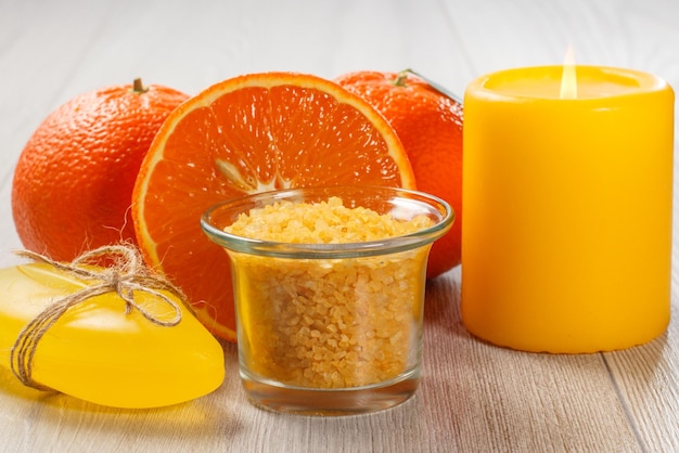 Couper l'orange avec deux oranges entières, un bol en verre avec du sel de mer jaune, du savon et une bougie allumée sur un bureau en bois. Produits et accessoires de spa