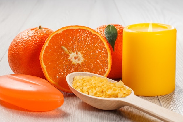 Couper l'orange avec deux oranges entières, du savon, une cuillère en bois avec du sel de mer jaune et une bougie jaune allumée sur un bureau en bois. Produits et accessoires de spa