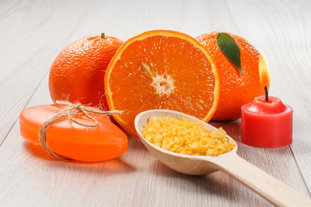 Couper l'orange avec deux oranges entières, du savon, une cuillère en bois avec du sel de mer jaune et une bougie rouge allumée sur un bureau en bois. Produits et accessoires de spa