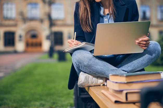Couper la photo d'une étudiante brune assise sur le banc avec des livres tout en écrivant à partir d'un ordinateur portable