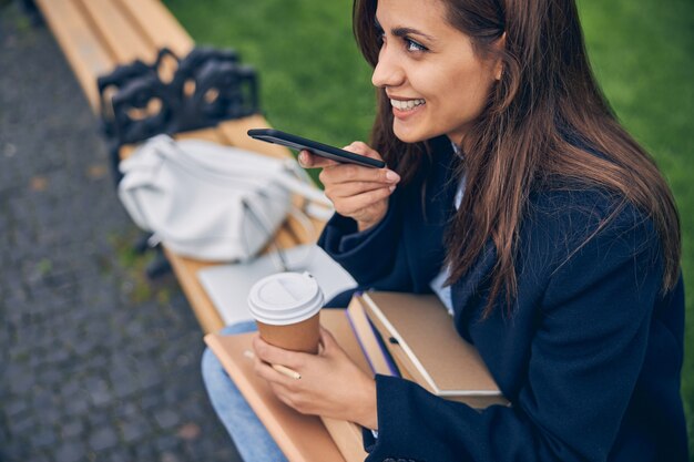 Couper la photo d'une jeune femme souriante assise sur le banc tout en parlant au téléphone