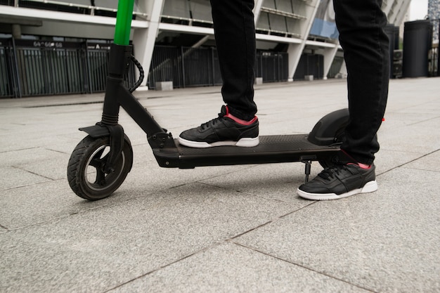 Couper la vue des jambes mâles debout sur l'e-scooter. Une façon intelligente de conduire dans une grande ville. Baskets élégantes noires. Guy a loué un scooter électrique par application smartphone. Habitudes écologiques.
