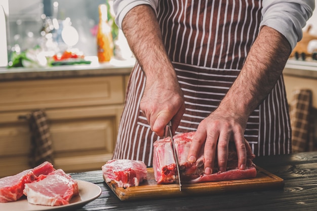 Coupes d'homme de morceau de boeuf frais sur une planche à découper en bois dans la cuisine à domicile. Un homme dans un tablier rayé avec un grand couteau dans ses mains