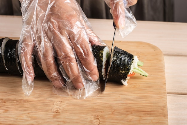 Photo coupez le kimbap (plat de riz en rouleau coréen) avec un couteau bleu. main de femme portant un gant en plastique, faisant gimbap, cut kimbap, korean roll gimbap (kimbob ou kimbap). faire du kimbop étape par étape