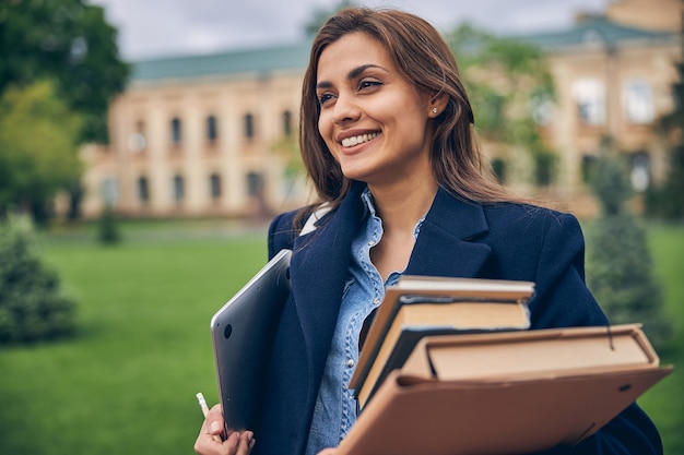 Coupez la photo d'un bel étudiant restant à l'extérieur près de l'université avec des livres et un ordinateur portable