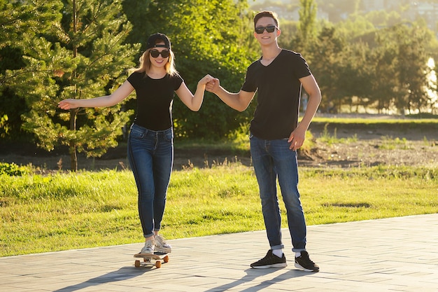 Couple actif amoureux du patinage sportif dans le parc d'été. une belle femme et un homme s'amusent avec un fond de planche à roulettes (longboard) la rivière. date de gars et de fille. mode de vie actif chez les jeunes