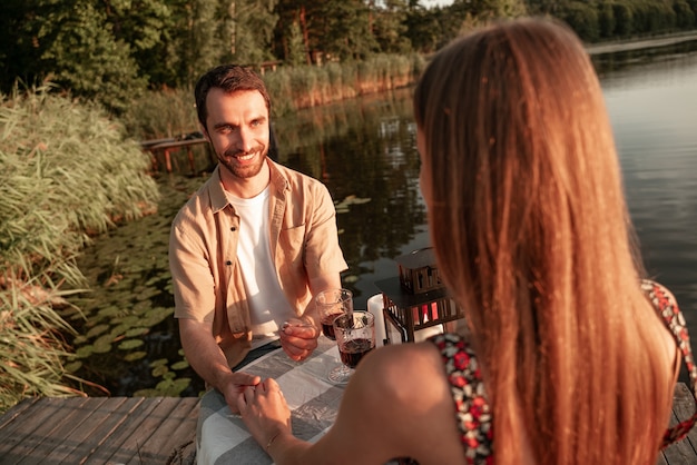 Un couple adorable passe du temps ensemble à boire du vin sur un pique-nique sur une jetée en bois : un bel homme tient la main de sa petite amie, fait une proposition, la regarde avec amour, le fond du lac forestier porte des vêtements d'été