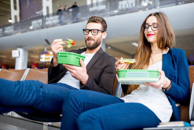 Couple d'affaires élégant mangeant avec des boîtes à lunch assis dans la salle d'attente de l'aéroport. Prendre une collation pendant un voyage d'affaires