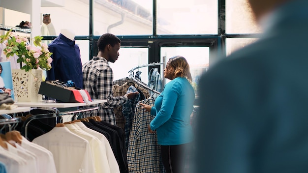 Couple afro-américain regardant des marchandises élégantes, vérifiant le tissu des vêtements dans un magasin de vêtements. Clients souriants appréciant une séance de shopping, achetant des tenues et des accessoires tendance dans une boutique moderne