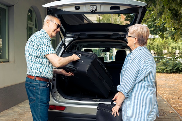 Un Couple âgé Met Des Sacs Dans Le Coffre Du Véhicule Pour Partir En Vacances à La Retraite Pendant L'été. Voyager En Voyage Sur La Route Pour Les Loisirs, Emballer Des Bagages Et Une Valise Pour Des Vacances Dans Le Paysage Urbain.