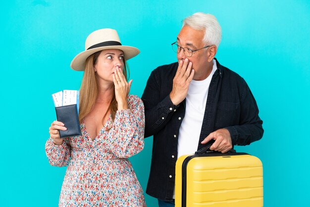 Couple d'âge moyen allant voyager et tenant une valise isolée sur fond bleu couvrant la bouche avec les mains pour avoir dit quelque chose d'inapproprié