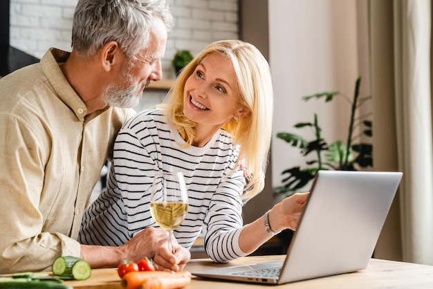 Couple d'âge moyen caucasien regardant un spectacle sur un ordinateur portable tout en cuisinant avec du vin à la cuisine