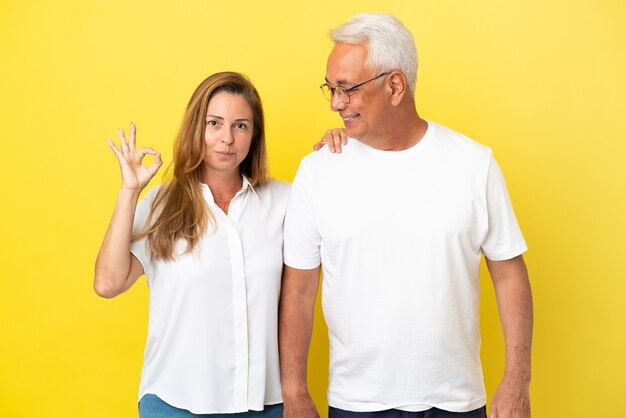 Couple d'âge moyen isolé sur fond jaune montrant un signe ok avec les doigts