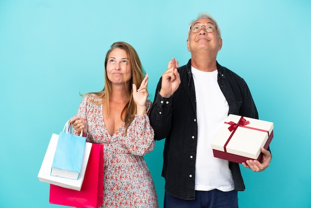 Couple d'âge moyen avec panier et cadeau isolé sur fond bleu avec les doigts croisés et souhaitant le meilleur