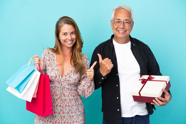 Couple d'âge moyen avec panier et cadeau isolé sur fond bleu donnant un geste du pouce avec les deux mains et souriant