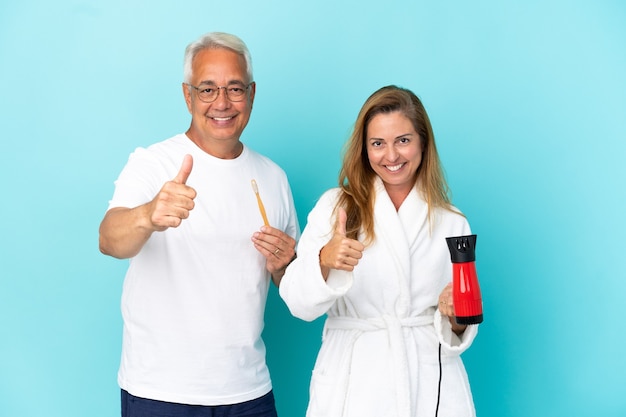 Couple d'âge moyen tenant une sécheuse et une brosse à dents isolées sur fond bleu donnant un geste du pouce parce que quelque chose de bien s'est produit