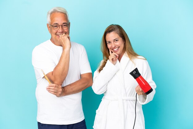 Couple d'âge moyen tenant une sécheuse et une brosse à dents isolées sur fond bleu souriant avec une douce expression