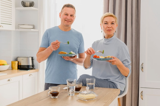 Couple d'âge mûr en bonne santé posant à la caméra tout en mangeant un petit déjeuner sain ensemble sur le milieu de la cuisine