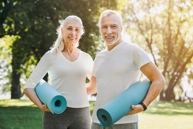 Couple d'âge mûr debout avec un tapis de yoga dans les mains dans le parc du matin avant l'entraînement avec regard sur la caméra